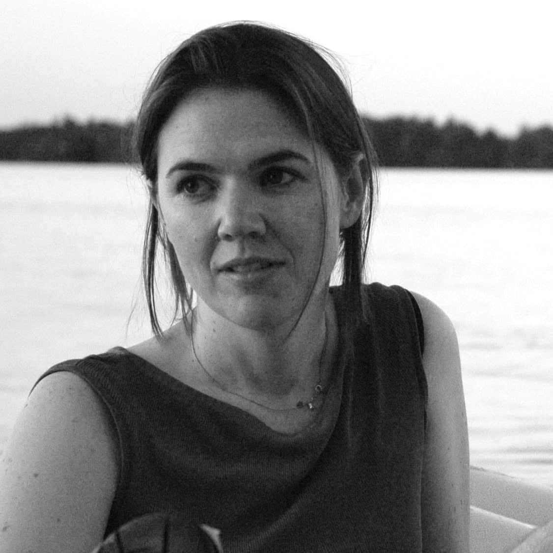 A person with long hair sits by the water, looking to the side, in a black and white photograph.