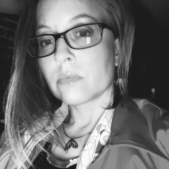 Black and white photo of a person wearing glasses, looking at the camera. They are wearing a jacket and a necklace with a pendant.