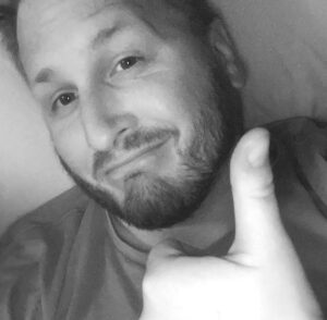 A person with a beard gives a thumbs-up, lying down, in a black and white photo.